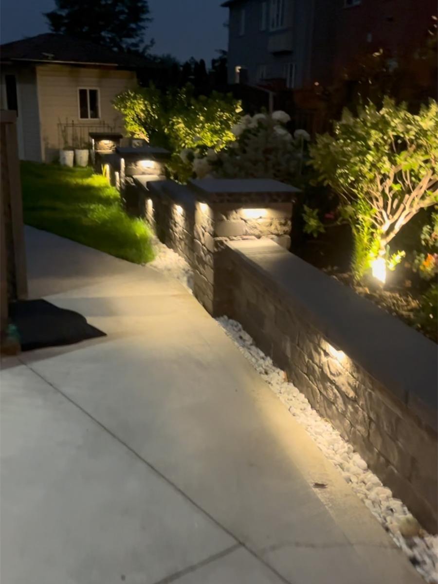Backyard at dusk with a lit stone retaining wall, garden lights and a wide walkway